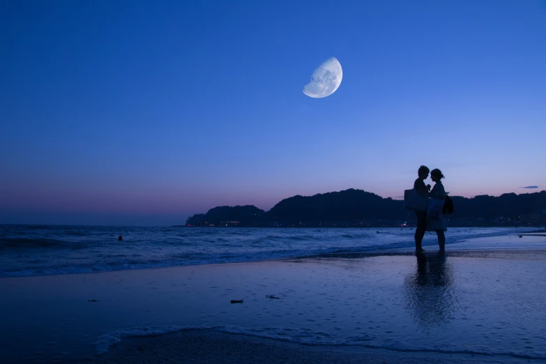 two people standing on top of a beach under a moon