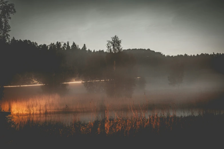 fog is shining on the night sky as the lights shine on a forest in front of a lake