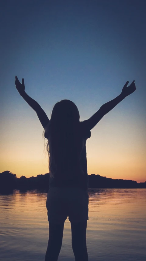 a person with their arms up standing by water at sunset