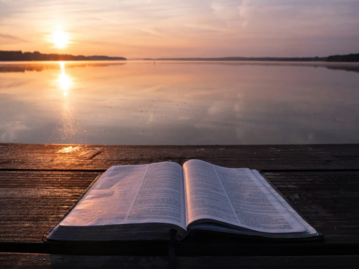 an open bible lies on a dock with the sun rising behind it