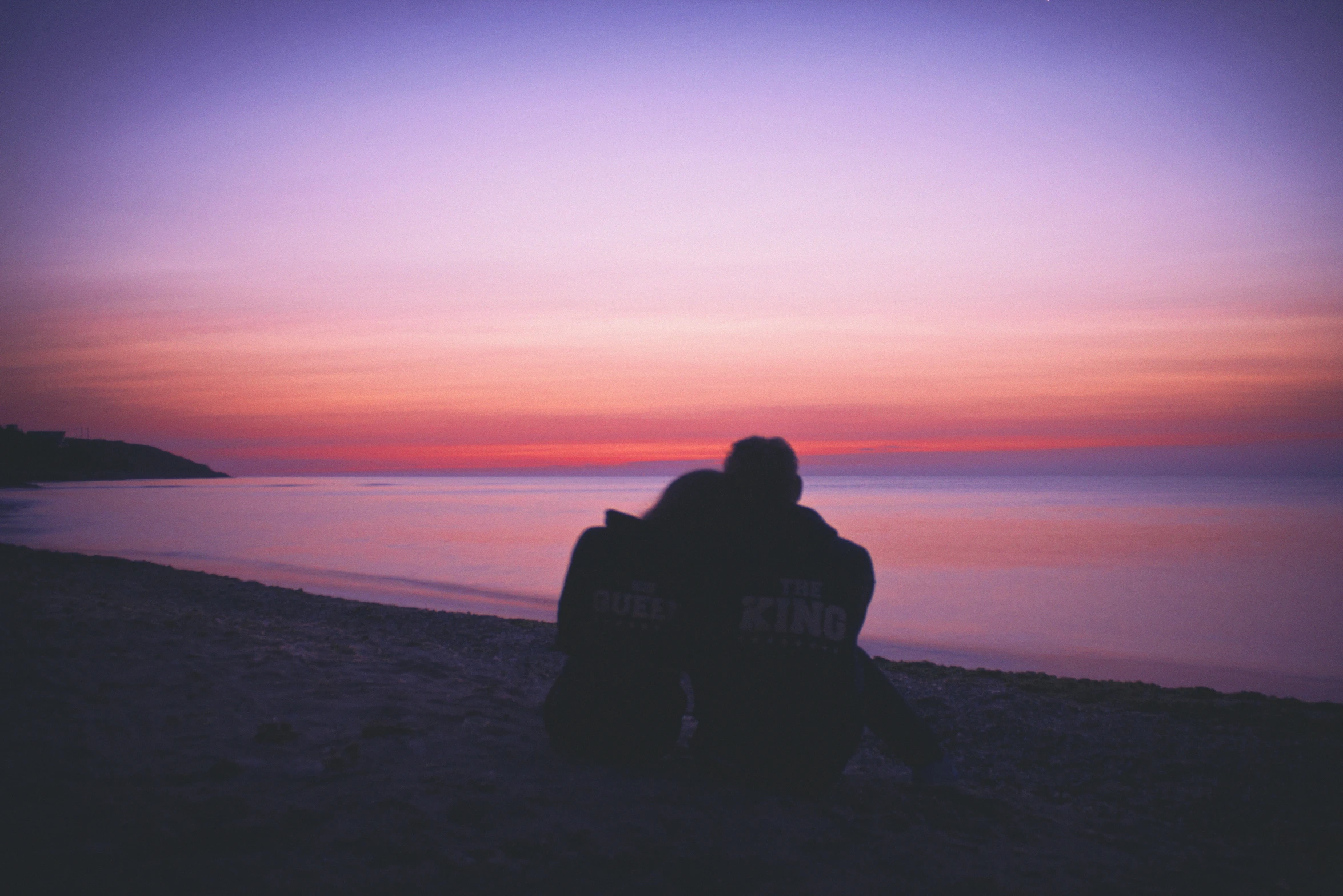 a couple sits on a bench next to the ocean at sunset