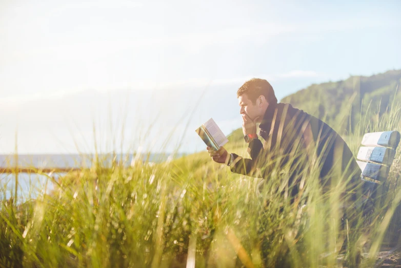 a man sitting on a bench reading a book in tall grass