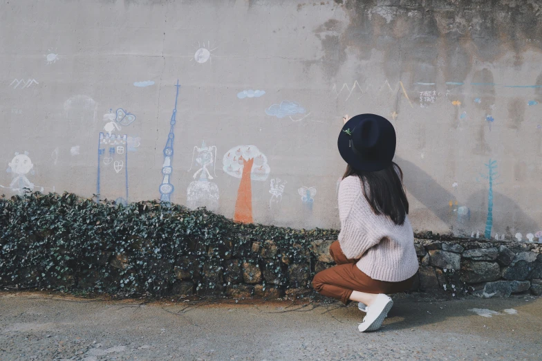a woman wearing white shoes and a black hat and squatting against a wall