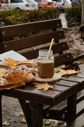 a glass of coffee, bagel and some leaves on a bench