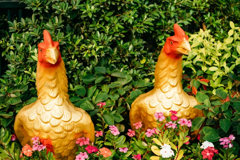 two golden rooster figurines sit in a garden surrounded by flowers