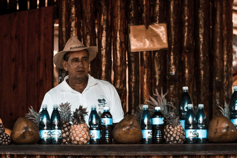 man with white shirt wearing straw hat selling pineapples and bottles