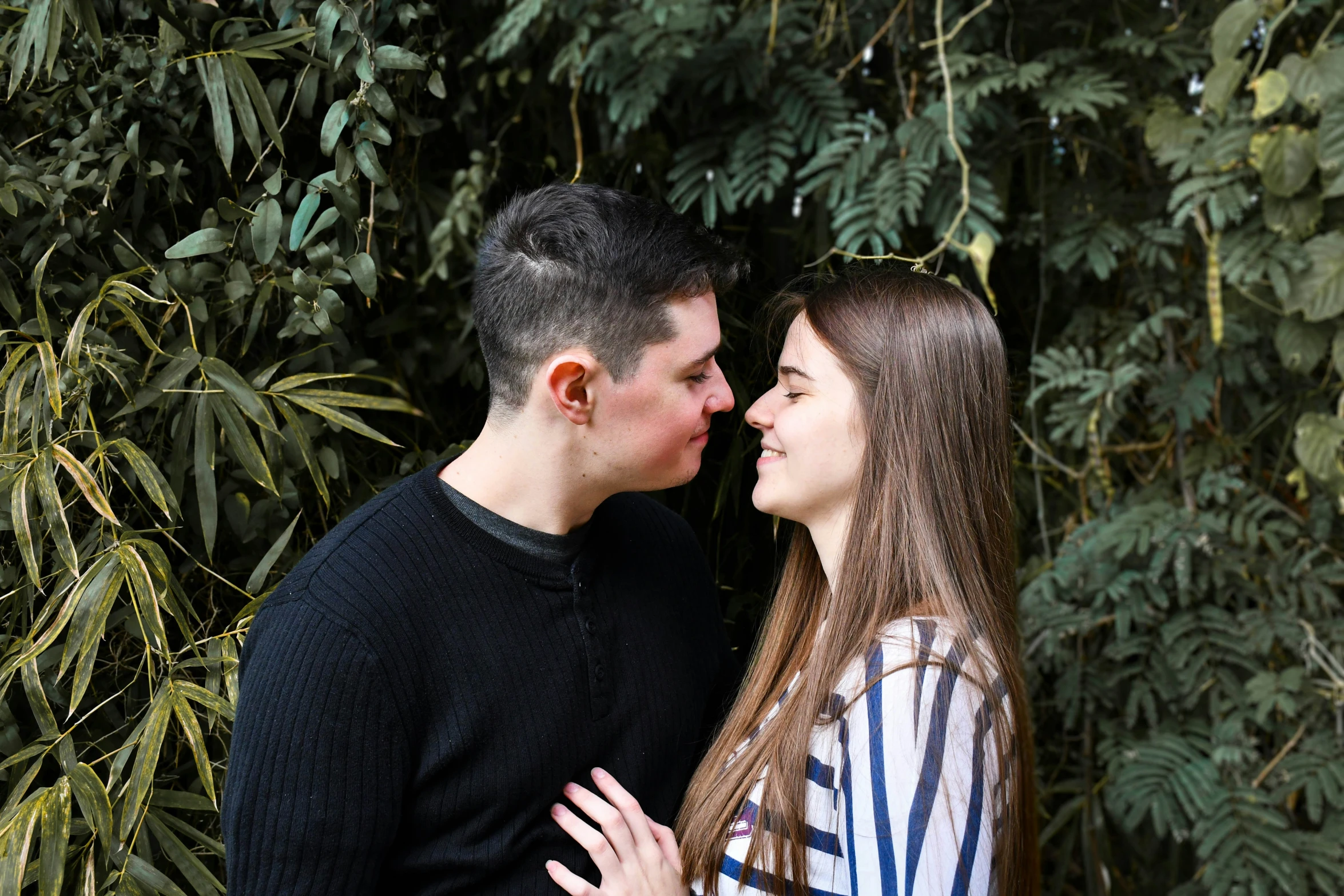 a beautiful young couple stands close together in front of a backdrop of greenery