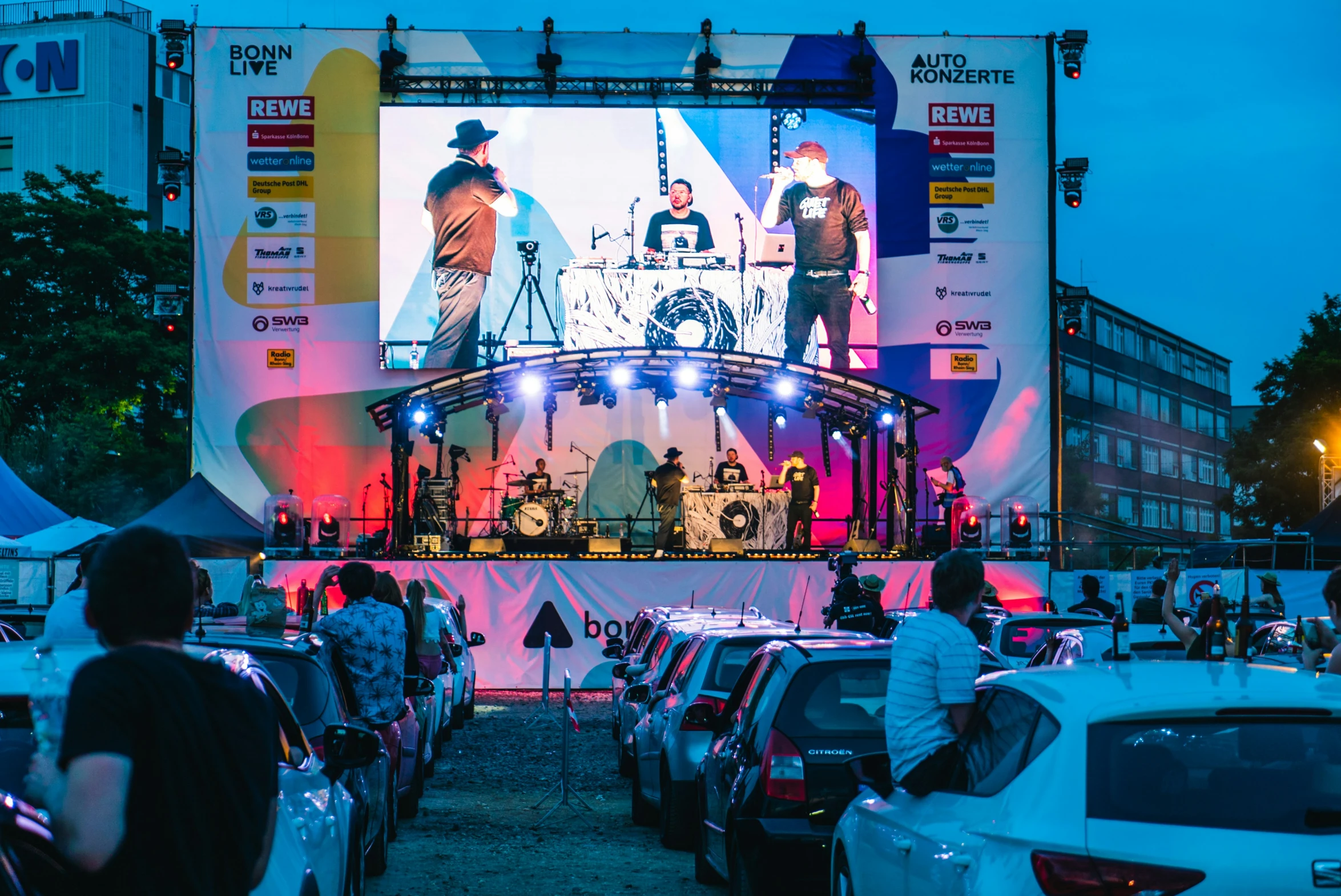 a giant screen featuring three musicians and a band plays an outdoor concert