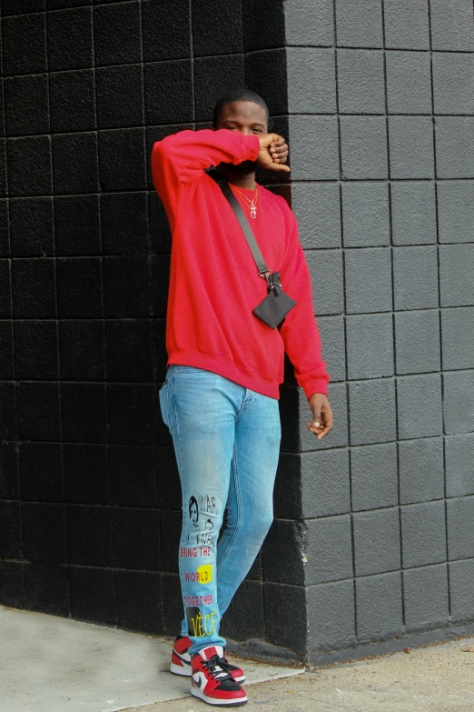 a man leaning on a brick wall while wearing blue jeans and a red sweater