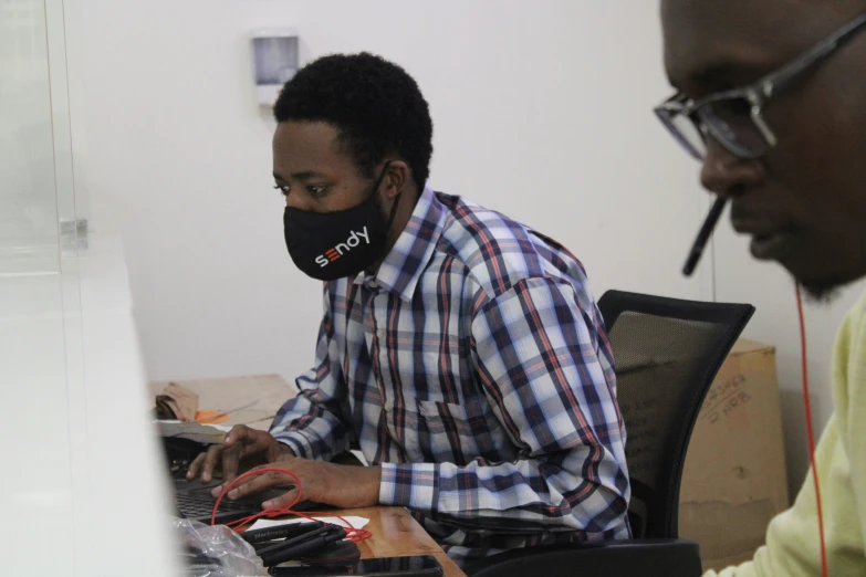 two people in the foreground and one is wearing a face mask while working on their computers