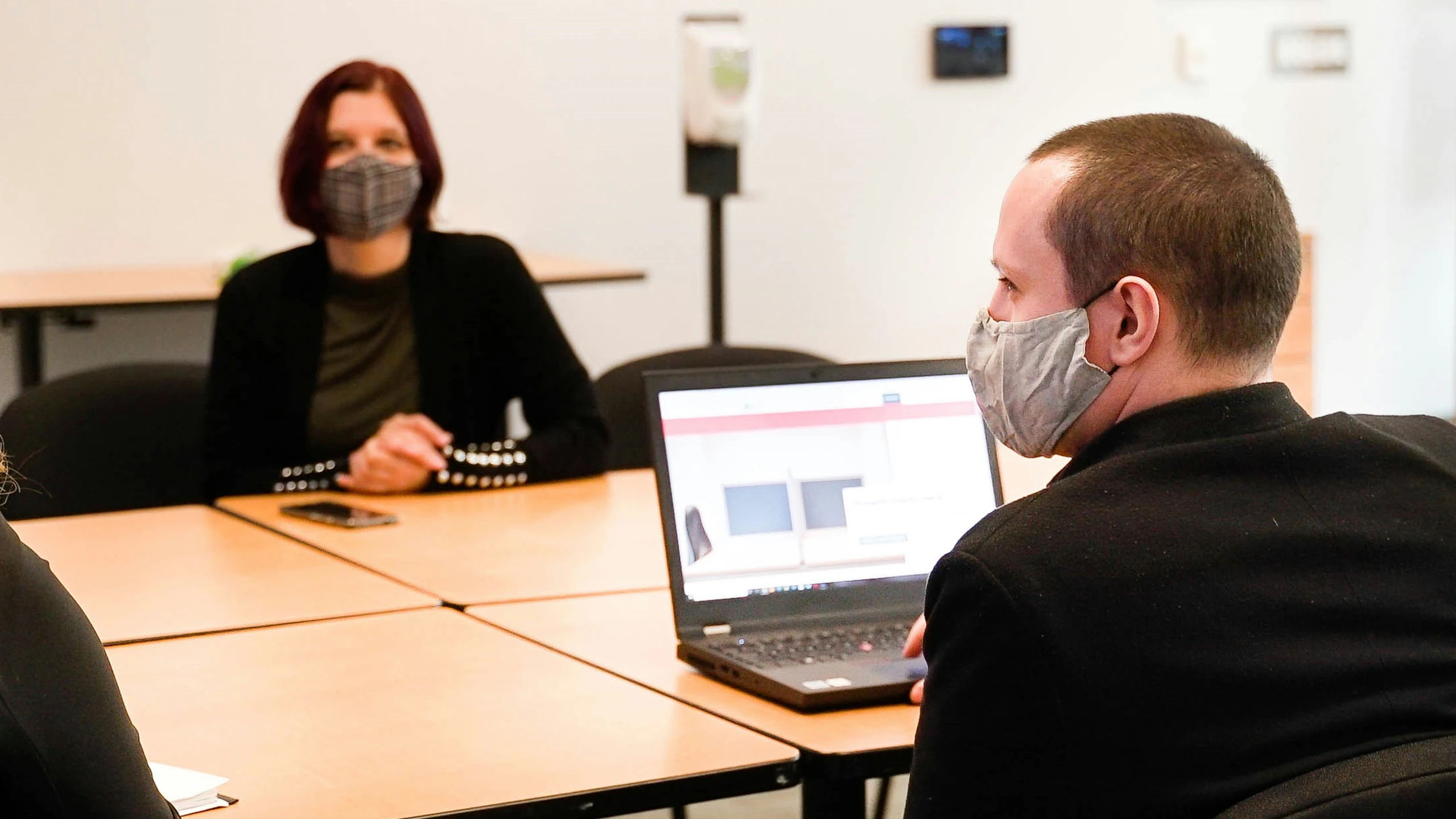 two people in masks sitting at a table with laptops