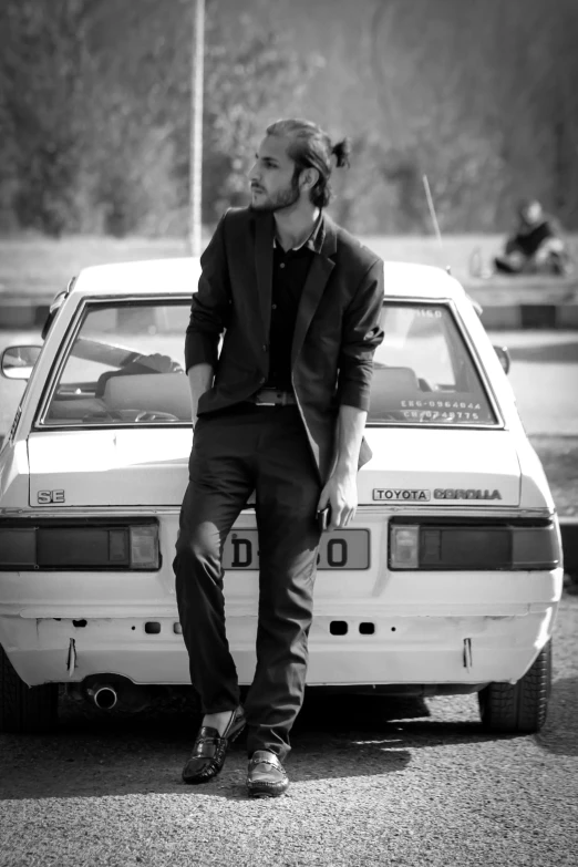 a man wearing a black suit and sneakers leaning against a car