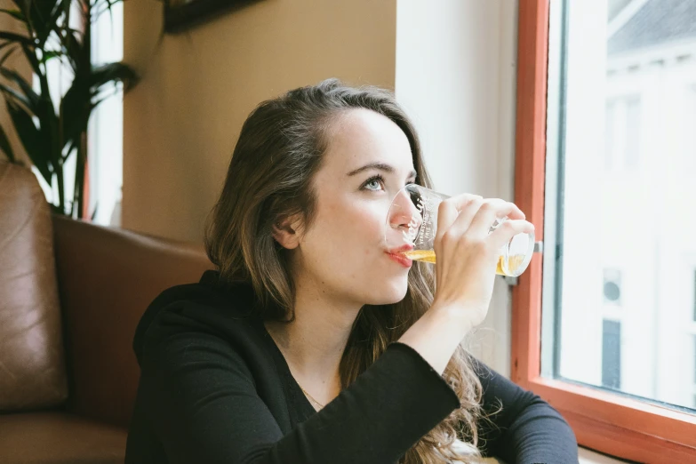 a girl drinking a drink from a glass sitting in a window sill