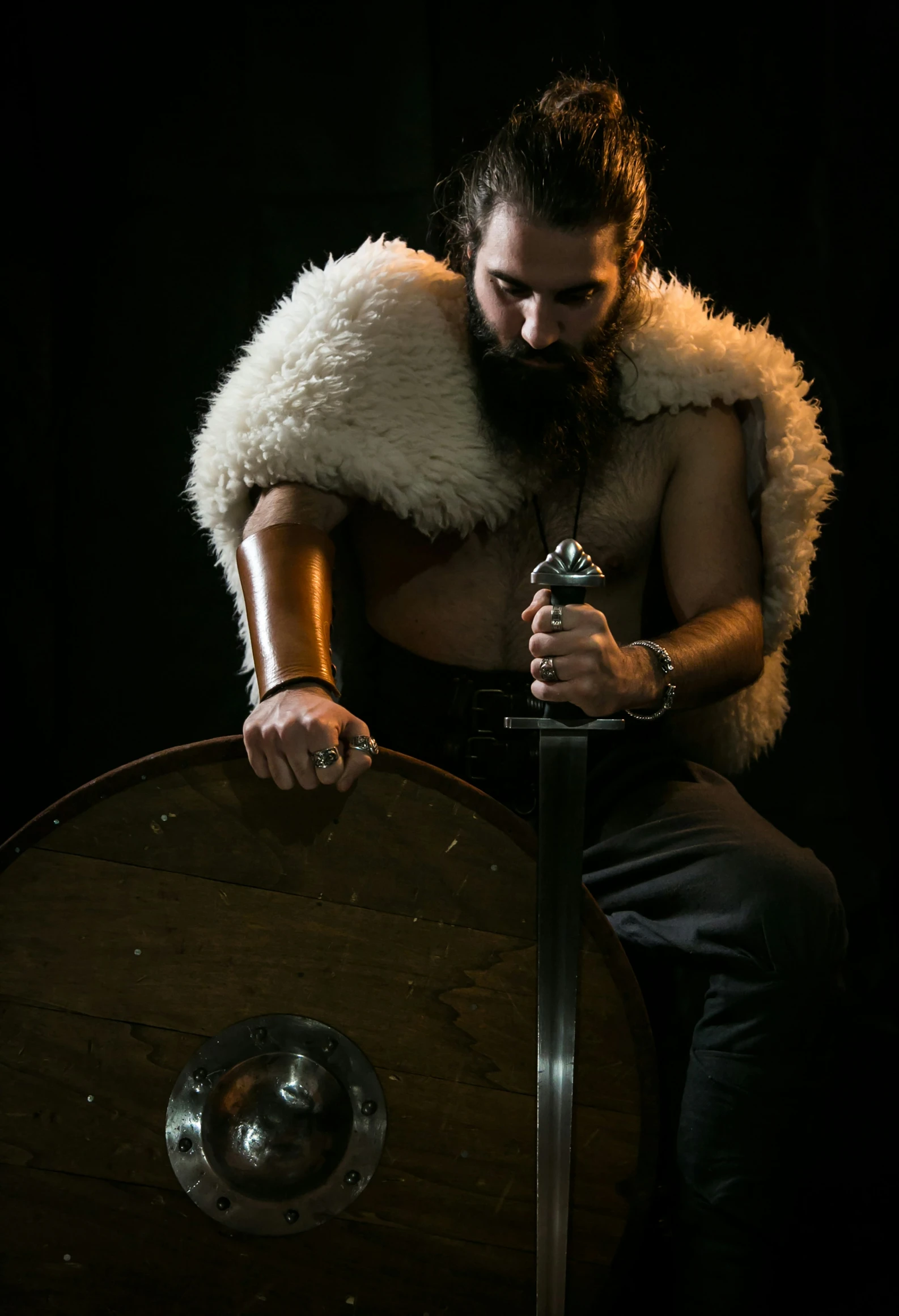 a bearded man dressed up with long hair and furs on his shoulders sitting near a sword