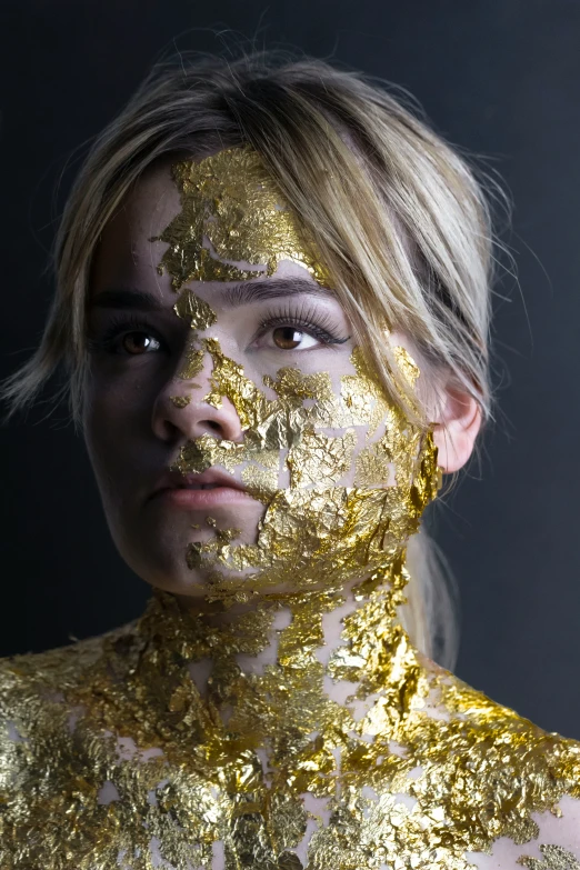 a young blonde woman has her gold painted face and body covered in gold leaf patterns