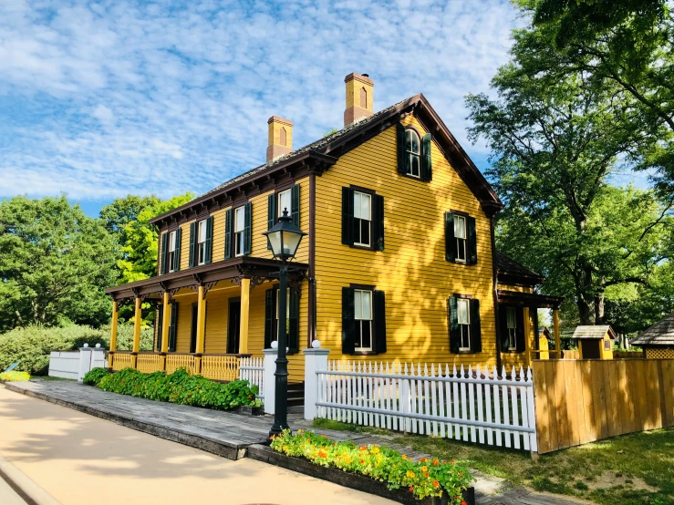 a tall yellow house has a white fence and some trees