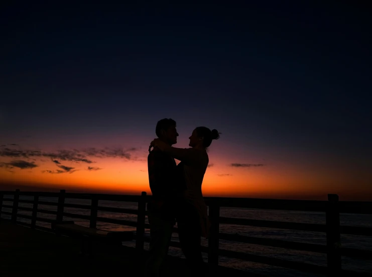 a couple standing at the end of a pier while the sun sets