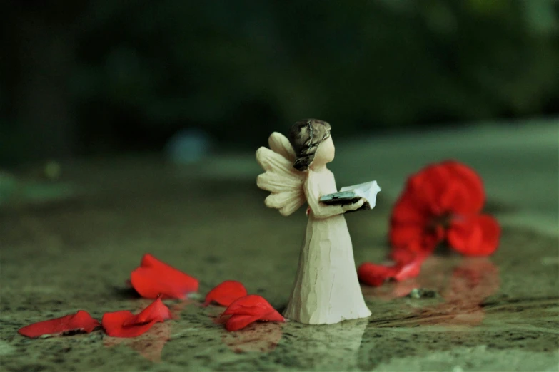 a small statue of a girl holding a magazine while a red rose petals are in the background