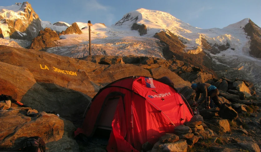 a red tent pitched on a rocky slope next to snow capped mountains