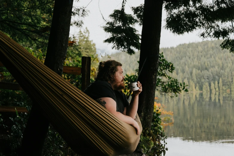 man sitting in hammock drinking coffee by the lake