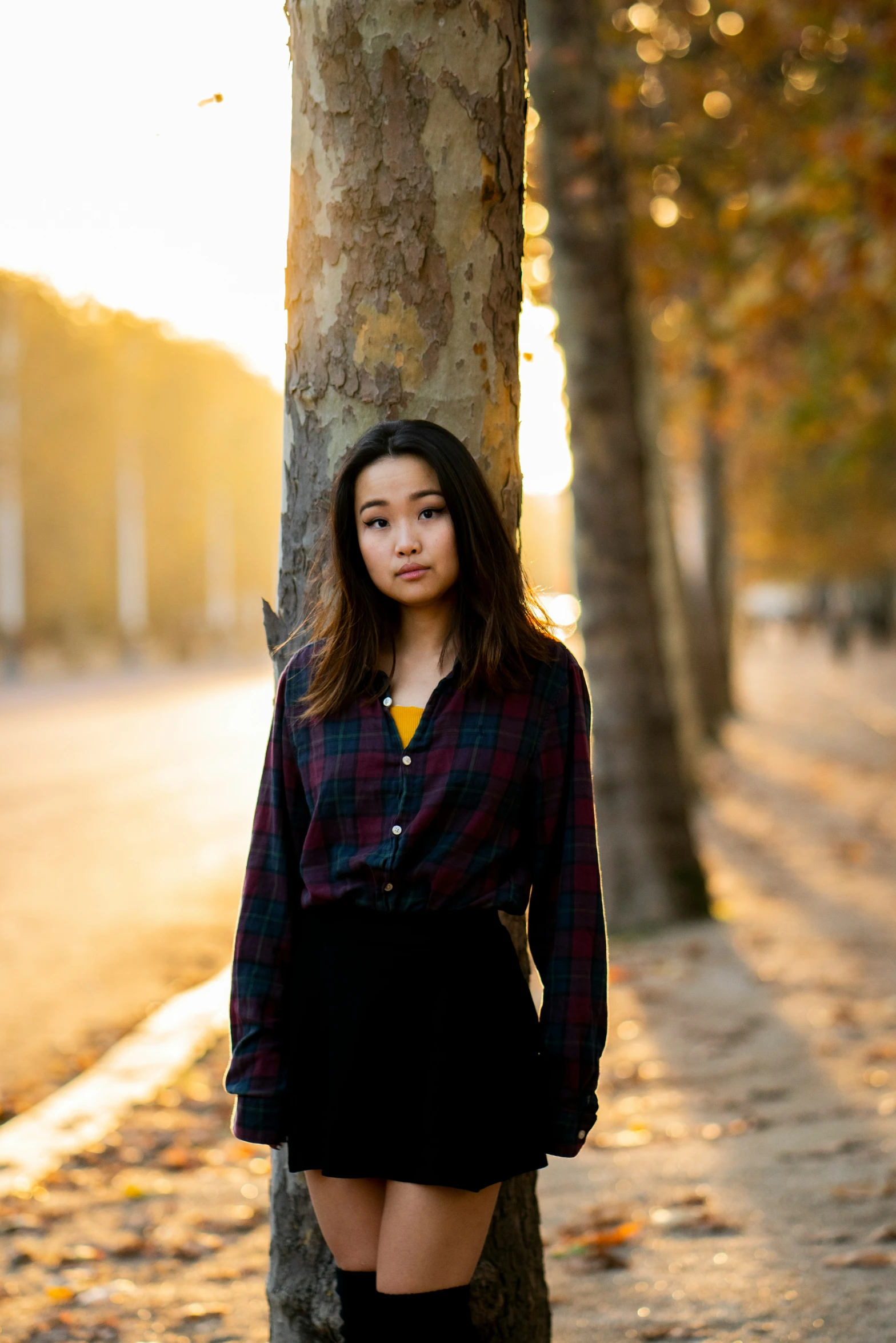 a girl stands next to a tree while wearing an oversize plaid shirt and black skirt