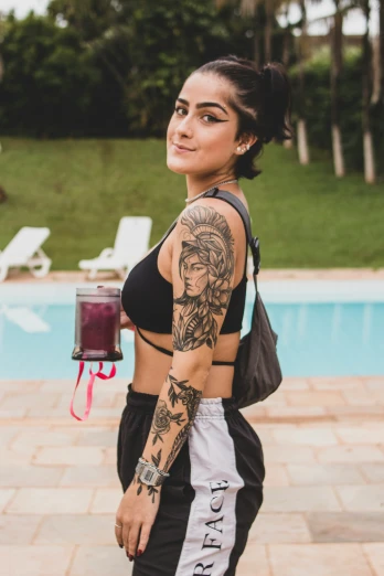 a woman wearing tattoos is holding a drink next to a swimming pool