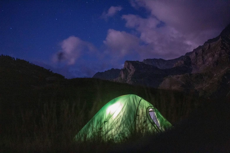 an illuminated green tent stands in the dark mountains at night