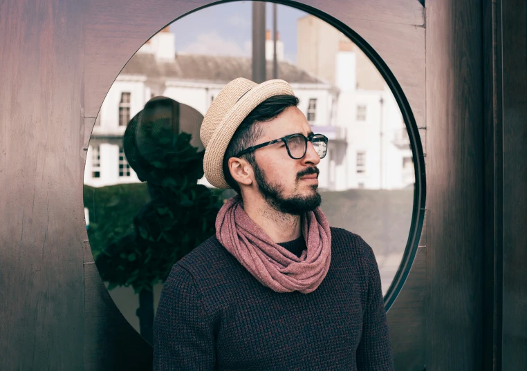 a man wearing glasses and a sweater is standing by a round mirror