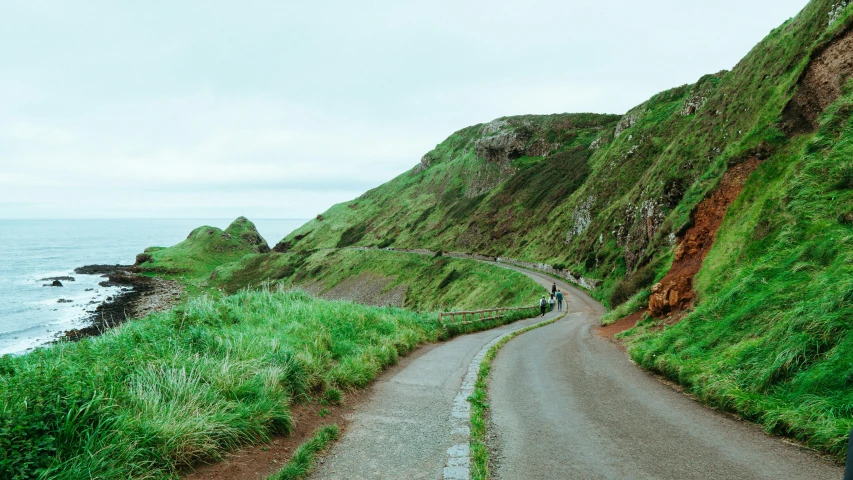 a man walks down a paved road with tall cliffs and water in the background