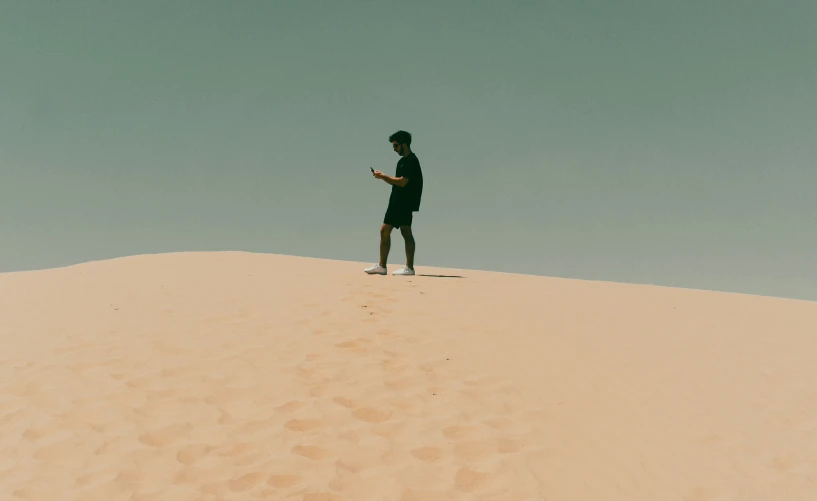 the man stands on top of a sand dune and looks at his cell phone