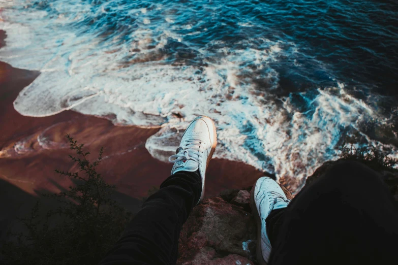 view from a cliff of a person's feet on the edge of the water and sandy shore