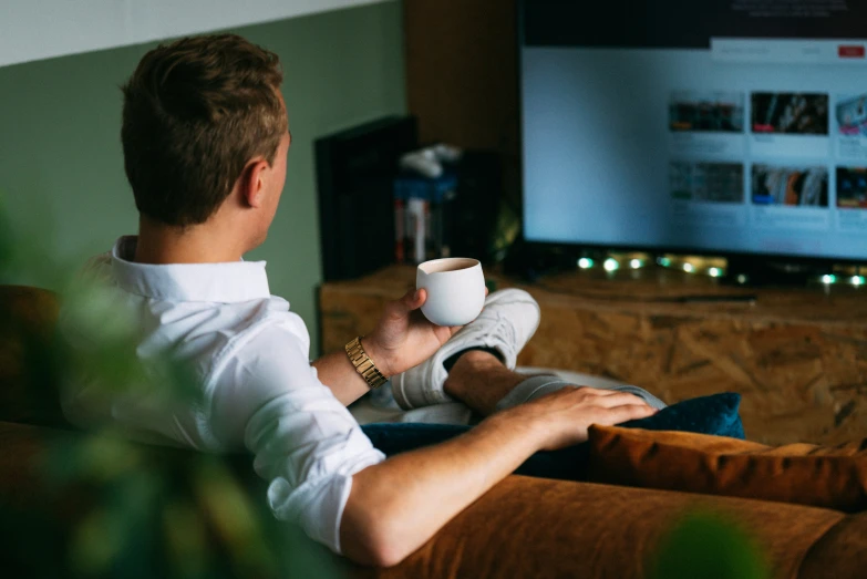 a person sitting on the sofa in front of the tv, holding up a cup of coffee