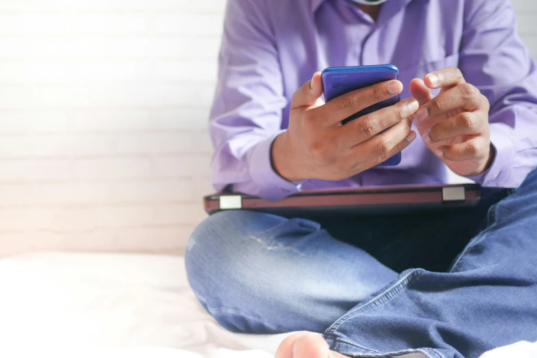 the person in blue is sitting on the bed and using his cell phone