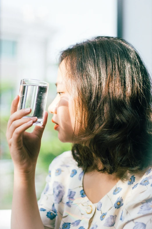 a woman sitting in front of a window, holding a glass up to her face