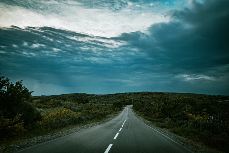 a long, empty road in a landscape with green grass and bushes on either side under cloudy skies