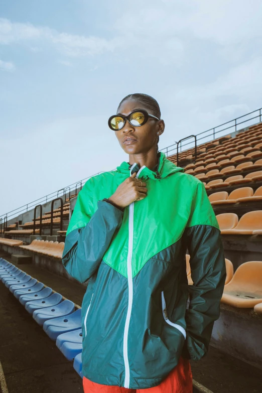 a man in sunglasses wearing an orange and green jacket and standing by some bleachers