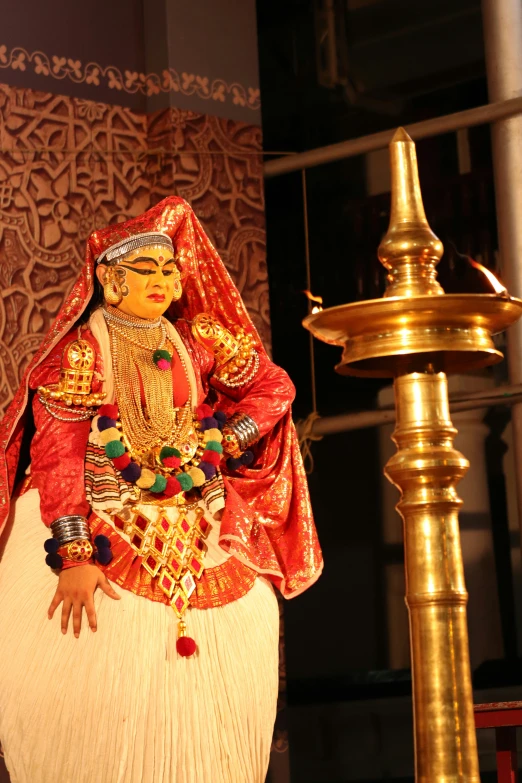 a woman wearing a colorfully dressed garb, with lots of gold ornaments, poses in front of a tall lamp