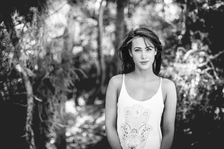 a woman in a white tank top standing near some trees