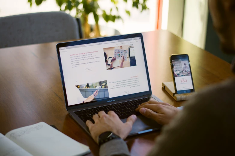 man sitting at table using laptop with cellphone, and book open