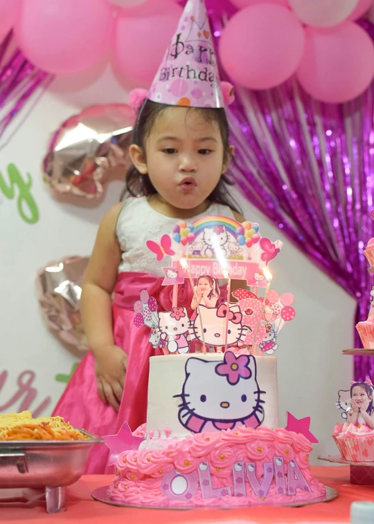 little girl dressed in pink is standing behind a hello kitty birthday cake