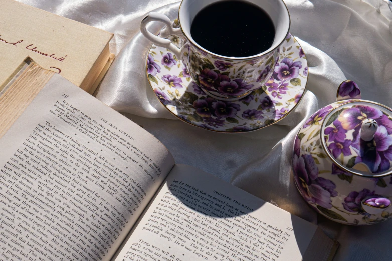 an open book on the table next to some tea cups and saucers