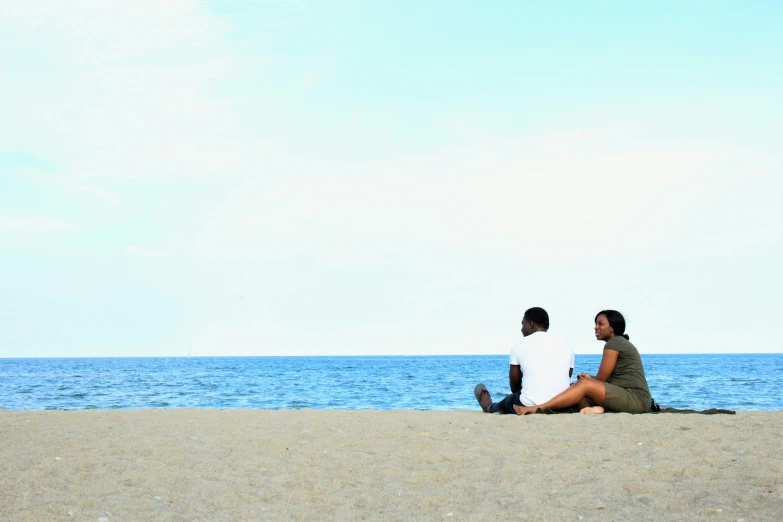 a man and woman are sitting on the sand at a beach, overlooking the ocean