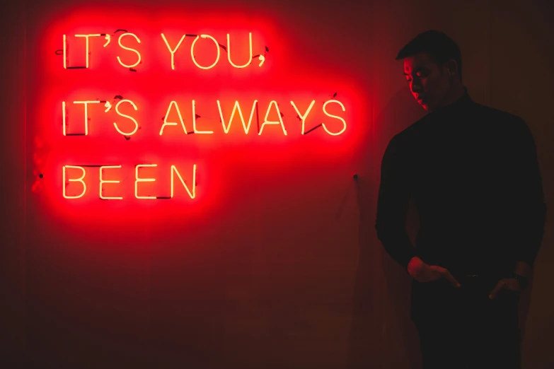 man standing in front of large neon sign that says it's you it's always been
