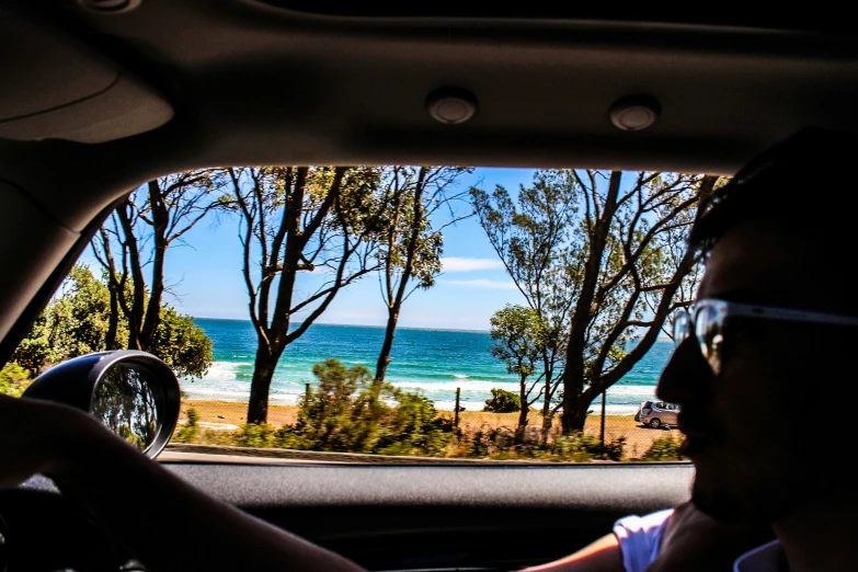 the inside of a car showing a man driving and the ocean in the back