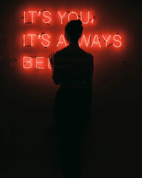 a woman stands in a dimly lit room, staring at a red neon sign
