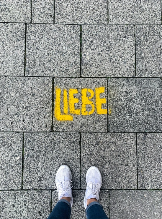 someone's feet with white sneakers on standing at the bottom of a yellow street sign