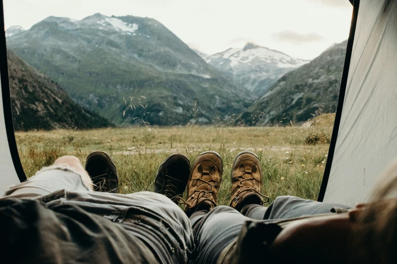 people in boots standing near a tent while laying in front of mountains