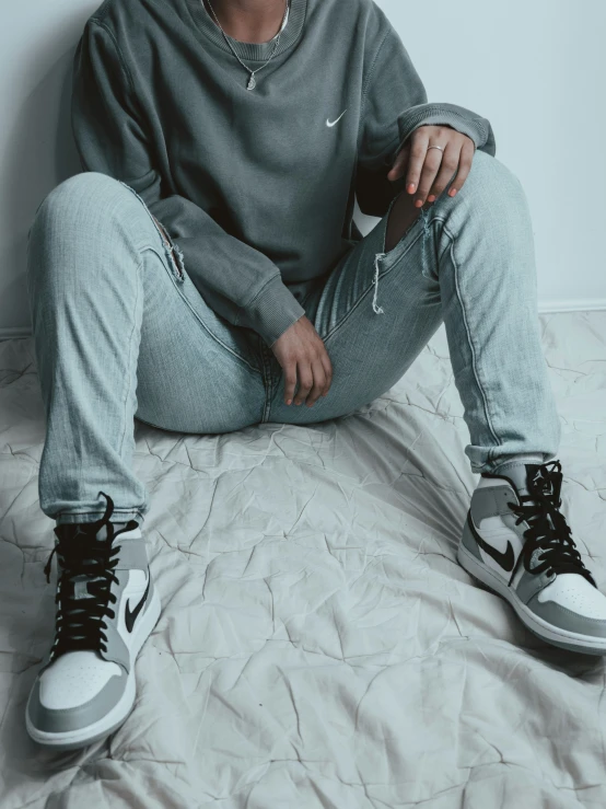 a person sits on a bed, wearing a grey sweatshirt and jeans