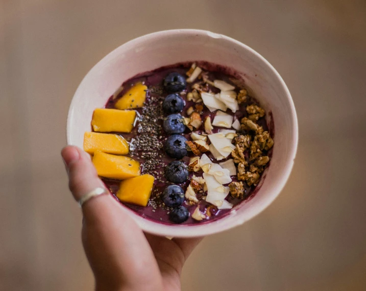 a hand holding a bowl filled with blueberries, mangoes, granola and oat