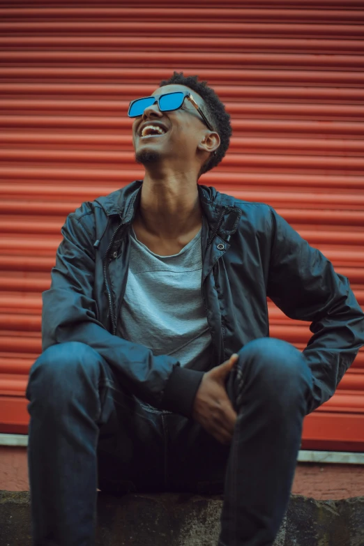 a young man sitting down laughing and wearing sunglasses